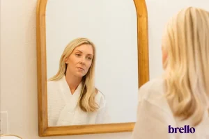 Woman practicing body acceptance while looking at her reflection in bathroom mirror