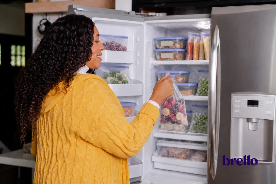 Woman holding a bag of frozen fruit beside an organized freezer stocked with prepared GLP-1 freezer meals and frozen vegetables.