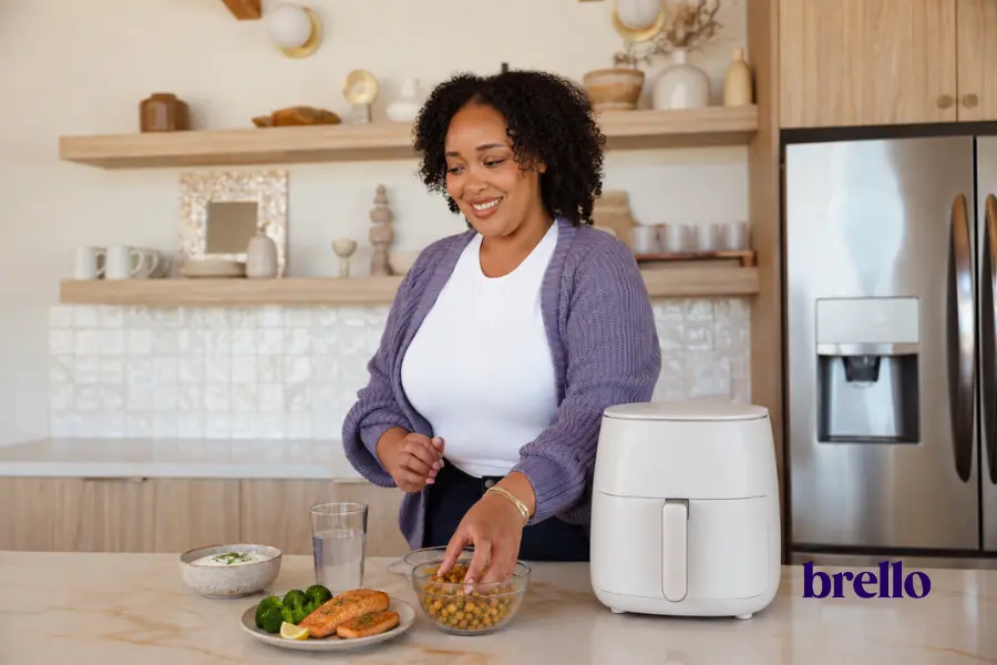 Woman tossing seasoned chickpeas at a kitchen counter beside an air fryer, with salmon and broccoli on a plate, example of GLP-1 air-fryer recipes.