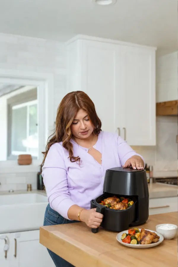 Woman opening an air fryer basket of chicken and vegetables at a kitchen counter